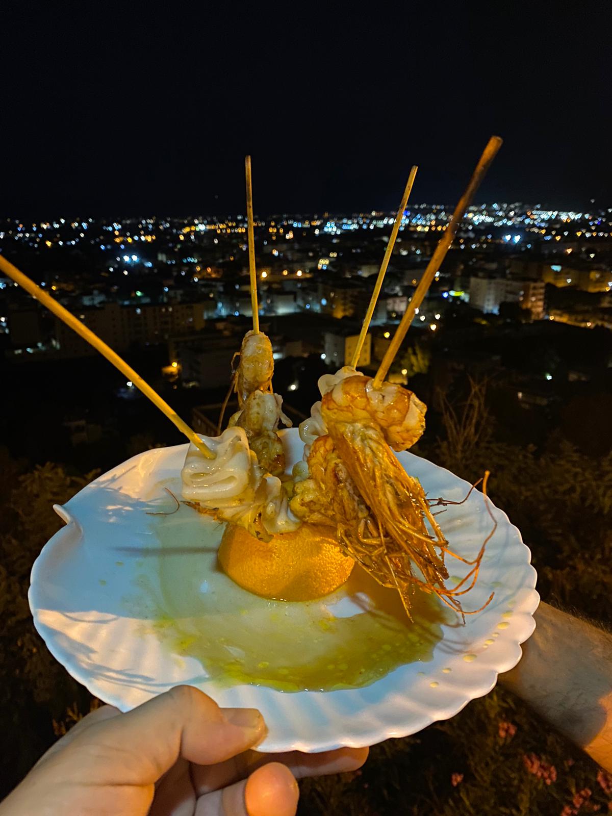 A hand holding a scalloped plate of grilled prawns and squid up against the lights of Carini at night